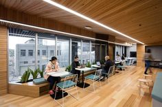 people are sitting at desks in an office with large windows and wooden floors, while one person works on a laptop