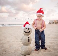 a baby boy standing next to a snowman on the beach wearing a santa hat