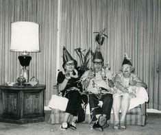 black and white photograph of three people wearing party hats sitting on a couch with drinks in their hands