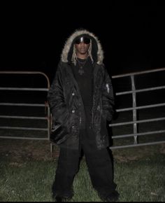 a man standing in the grass wearing a black jacket and sunglasses with a hood on