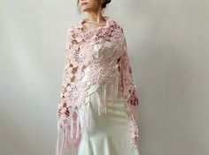 a woman wearing a pink crochet shawl and white dress standing in front of a wall