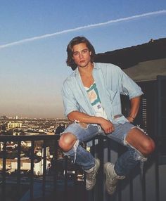 a young man sitting on top of a balcony next to a railing with the city in the background