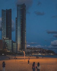 several people are on the beach at night near some tall buildings in the distance,