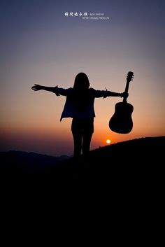 a woman holding an acoustic guitar while standing on top of a hill with her arms outstretched