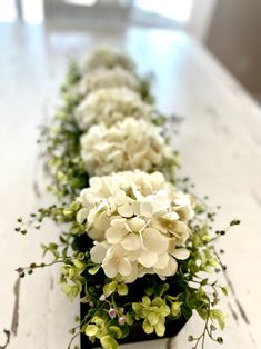 there is a long row of white flowers on the table with greenery in it