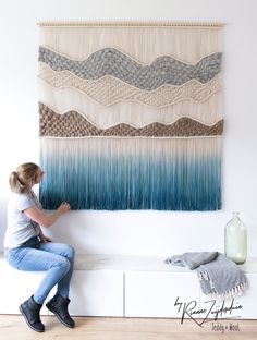 a woman sitting on top of a white bench next to a wall hanging with fringes