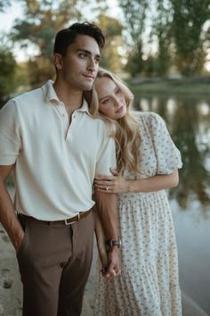 a man and woman standing next to each other near the water with trees in the background