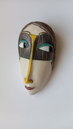 a white mask with blue eyes and yellow lines painted on it's face, against a white background