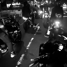 several people riding motorcycles on a city street at night with their lights on and one person standing next to them