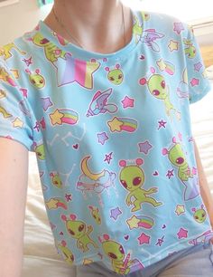 A pastel blue alien shirt featuring little green aliens who are only out to abduct your heart and bring a little shock of pastel to your life. This cropped t-shirt, which has a slightly silky, knit feel, also has contrasting rainbows and stars. It's unisex and fits loose, making for a comfy casual addition to your fairy kei and decora wardrobe while being low-key. Apparel and bedding ship separate from handmade items. Delivered in 2-4 weeks. The colors print more pastel than the items I make mys Cute Short Sleeve T-shirt With Star Print, Kawaii Crew Neck Top With Funny Print, Kawaii Funny Print Crew Neck Top, Cute Light Blue T-shirt With Cartoon Print, Kawaii Blue Tops For Summer, Blue Kawaii Tops For Summer, Multicolor Short Sleeve Kawaii Tops, Blue Kawaii Top For Summer, Blue Kawaii Summer Tops