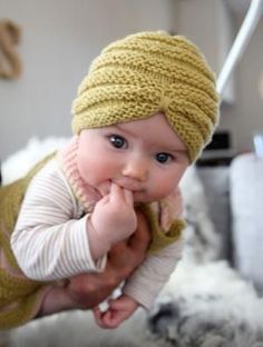 a baby wearing a yellow knitted hat and holding it's hand up to its mouth