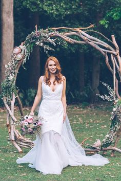 a woman standing in front of a wooden arch with branches and flowers on it, wearing a wedding dress