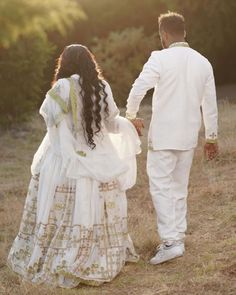 a man and woman dressed in white are walking through the grass holding each other's hands