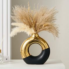 a gold vase with dried grass in it on a white table next to a candle