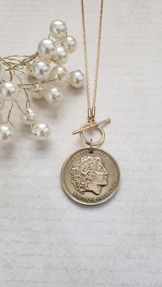 This authentic and vintage Greek coin makes a fabulous gold medallion necklace. It is a  1994 100 drachma coin set on an 18" gold-plated chain. Your choice of a front toggle closure or a back lobster clasp with a 3" chain extender. The coin has been polished and matches the chain beautifully. Welcome to Recycled Finery!  I repurpose old jewelry, coins, beads, vintage buttons, watch parts, and charms into new jewelry.  My ever-growing collection of these materials comes from estate sales, rummage sales, antique and thrift stores, and family and friends' collections they no longer need or want. I have fun putting these new pieces together and hope you will find them fun to wear and as unique gifts for your loved ones.     Please note: Because my components are recycled from old or vintage je Gold Coin Necklace Nickel Free, Nickel-free Yellow Gold Medallion Coin Necklace, Classic Gold Medallion Necklace With Coin Pendant, Antique Gold Coin Necklace Nickel Free, Antique Coin Necklace As A Gift, Vintage Charm Coin Necklace With Round Pendant As Gift, Medallion Coin Necklace With Vintage Charm As A Gift, Commemoration Necklace With Coin Pendant, Antique Coin Necklace For Gift