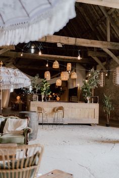 This shows a wedding bar display in a rustic barn. The bar display features cane bar stools, live trees, wooden crates and vases of dried flowers. Above the bar are cane lampshades with warm light bulbs. Live Tree