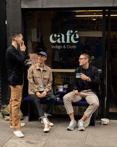 three men are sitting on a bench outside cafe indigo and cloth, talking to each other