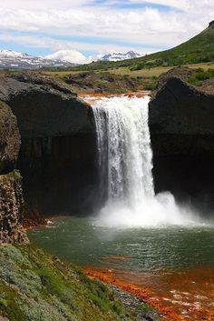 a waterfall with red algae in the water and green grass on the ground next to it