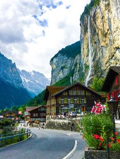 the road is lined with wooden buildings and flowers in front of tall mountain peaks that rise high into the sky