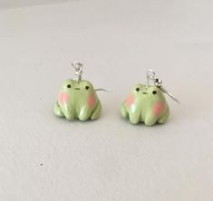 two small green and pink frog earrings with silver hooks on white table top, one is wearing