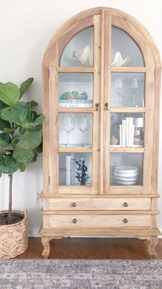 a wooden china cabinet with glass doors and shelves on the top, next to a potted plant