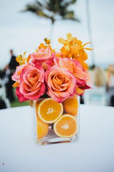 an arrangement of oranges and roses in a square vase on a table with other flowers