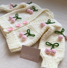 two knitted sweaters with pink flowers and green bows on the front, one has a name tag attached to it