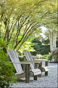 two wooden chairs sitting next to each other on top of a gravel road near trees