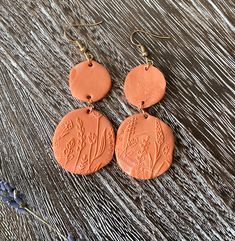 two orange earrings sitting on top of a wooden table next to purple flowers and leaves