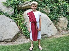 a young boy dressed in roman garb standing next to large rocks