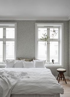 a white bed sitting under two windows in a bedroom next to a wooden table and stool