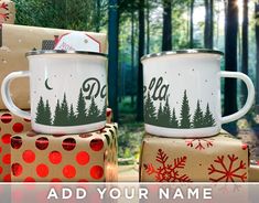 two white coffee mugs sitting on top of christmas presents in front of the woods