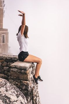 a woman sitting on the edge of a cliff with her arms up in the air