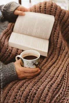 a person holding a cup of coffee and an open book on top of a blanket