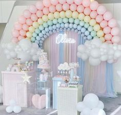 a table topped with balloons and cake next to a rainbow shaped arch covered in pastel colors