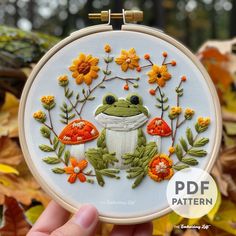 a hand embroidered frog with flowers and leaves on it's face, sitting in front of a leafy background