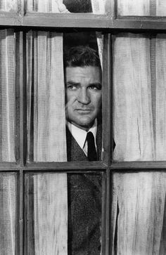 black and white photograph of man looking out window with curtains behind him, in front of bookshelves