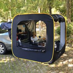 a car is parked next to a tent in the woods with its door open and two people sitting inside