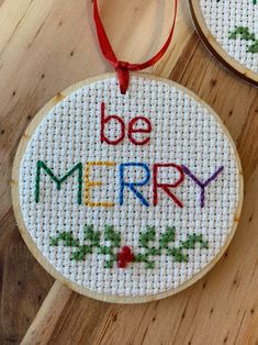 two cross - stitch christmas ornaments on a wooden surface, one with the words be merry