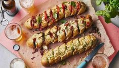 two long sandwiches are on a cutting board next to beer glasses and utensils