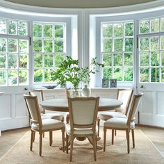 a dining room table with four chairs and a vase on the table in front of three windows