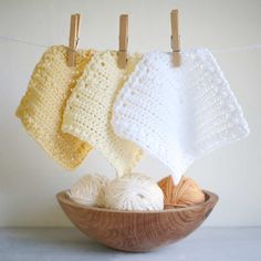 three crocheted dishcloths hanging on clothes pins in a bowl with yarn balls