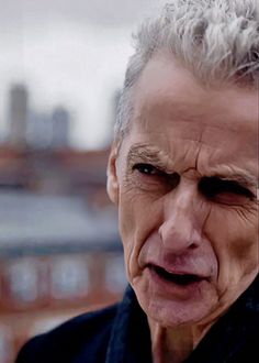 an older man with grey hair and black shirt looking at the camera while standing in front of a cityscape