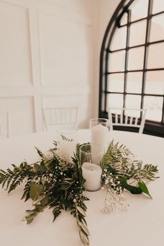 candles and greenery are arranged on the table in front of an arched glass window