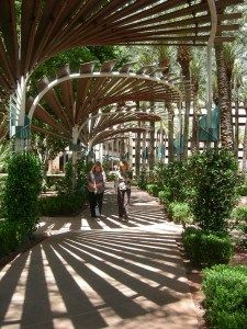 two people are walking down the sidewalk under an umbrella covered walkway that is lined with trees and bushes