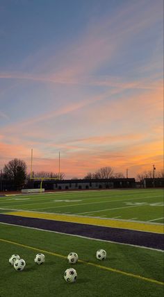 several soccer balls sitting on the grass in front of an empty football field at sunset