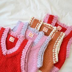 five knitted sweaters are lined up on a white sheet, one is red and the other is pink