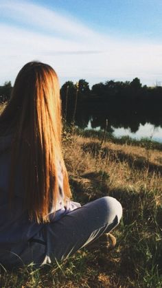 a woman sitting in the grass looking out over a body of water on a sunny day