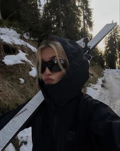 a woman in sunglasses holding a snowboard on the side of a snowy mountain road