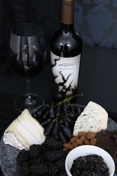 a wine glass and some cheese on a plate with blackberries, nuts and crackers